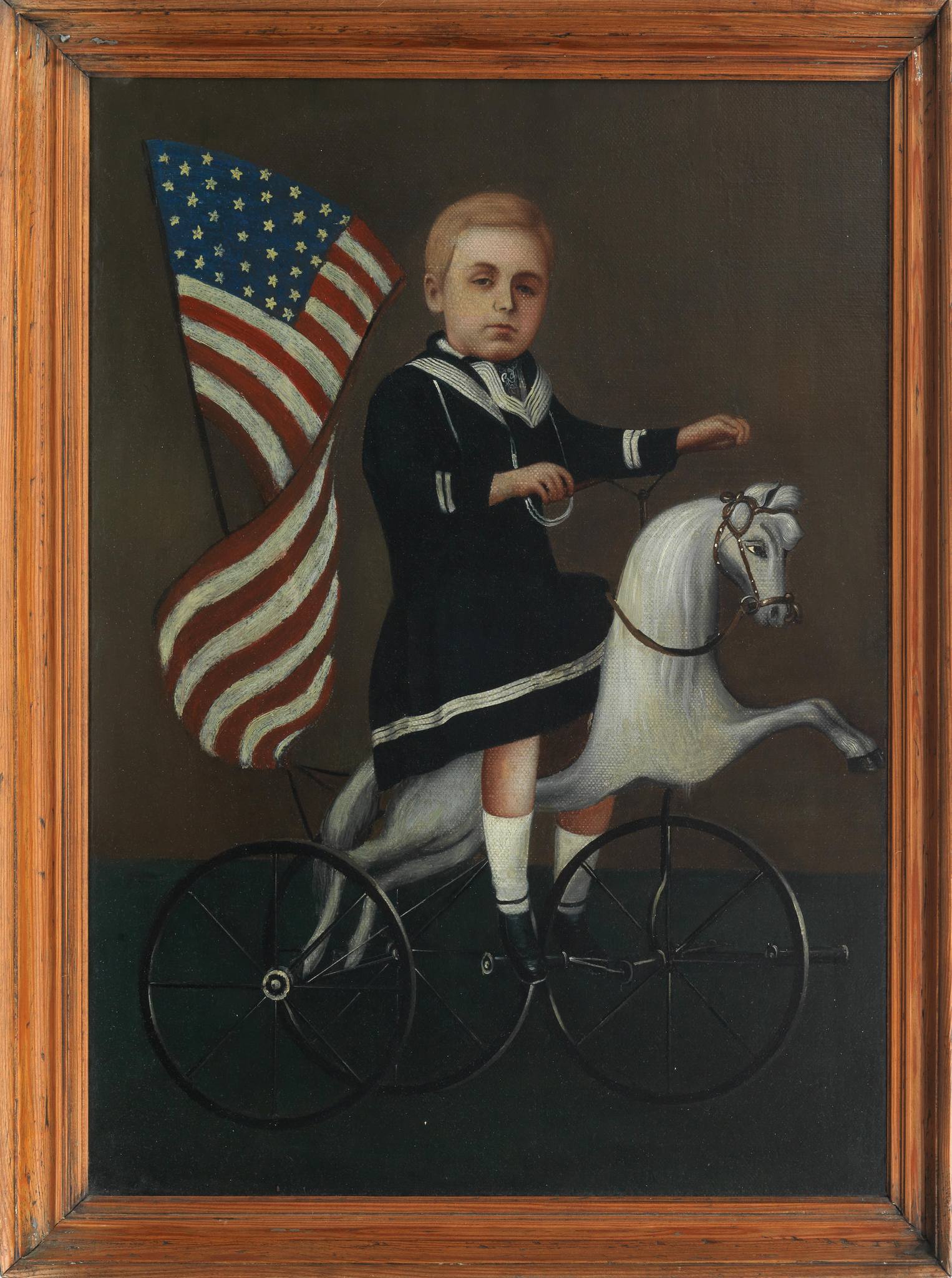 Portrait of a boy on a velocipede horse tricycle, with a large American flag waving in the background.